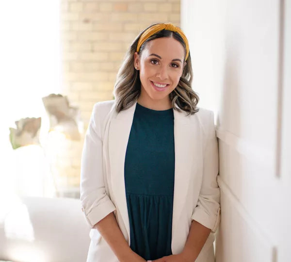 Stephanie Muskat, founder of Compassion in Caregiving, wearing a white blazer and dark green blouse, leaning against a white wall